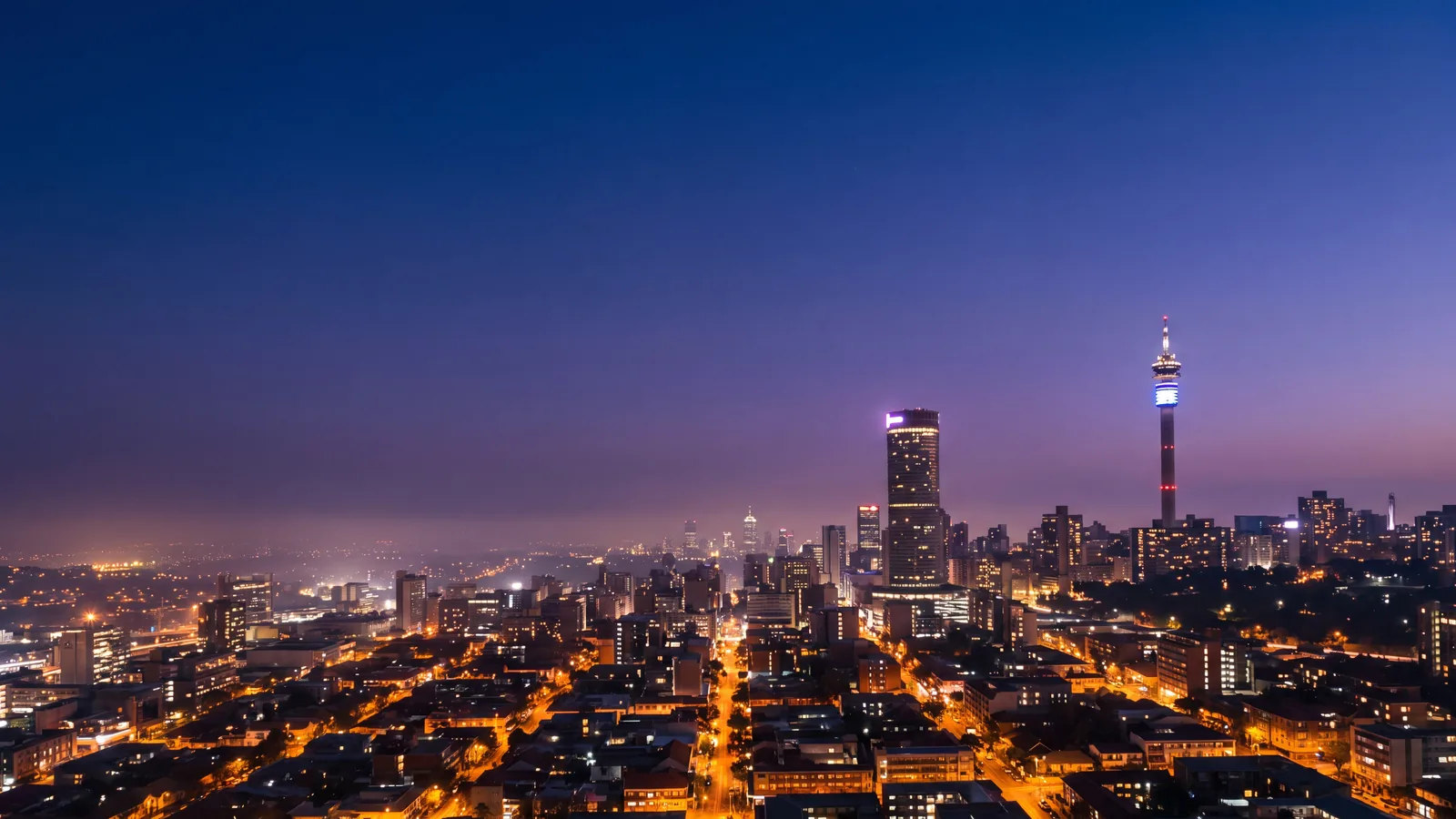 The Johannesburg skyline at blue hour with the Hillbrow tower and the city grid glowing amber under a violet sky